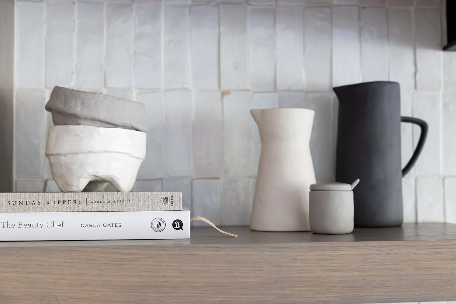 Ceramic vessels and cookbooks on an oak shelf against white tile. Devotion, Ancala, Desert Dwelling.