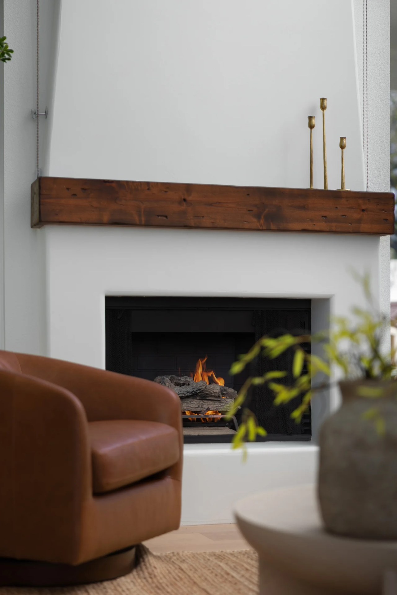 A white plaster fireplace with a walnut beam, brass candlesticks, and a leather chair. New Beginnings, Scottsdale, Desert Dwelling.
