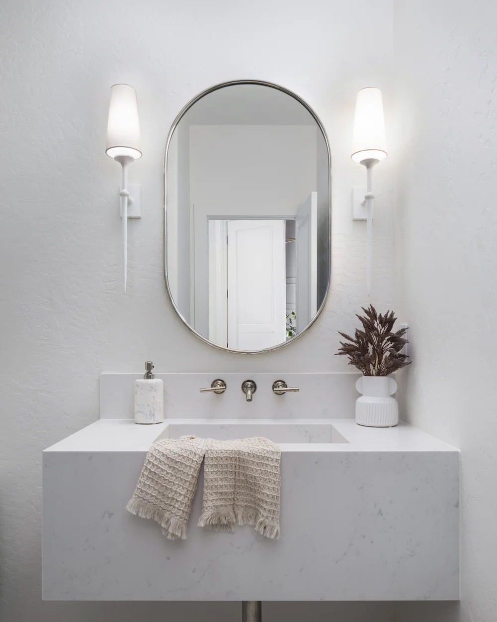 A powder room with a floating marble sink, an oval mirror, and white sconces. Tuckey, Scottsdale, Desert Dwelling.
